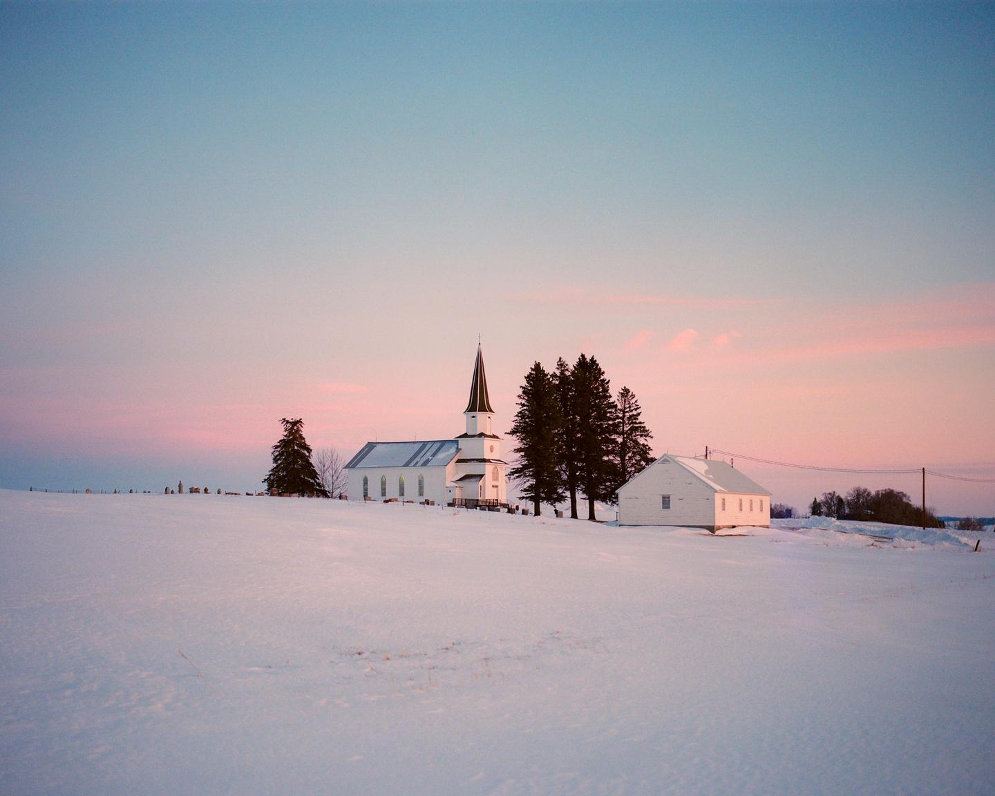 A white church with a tall steeple stands in a snow-covered field next to a small white building and several evergreen trees under a pink and blue sky.