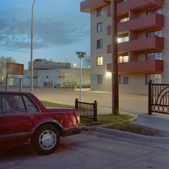 A red car is parked on a street next to an apartment building with a "no vacancy" sign.
