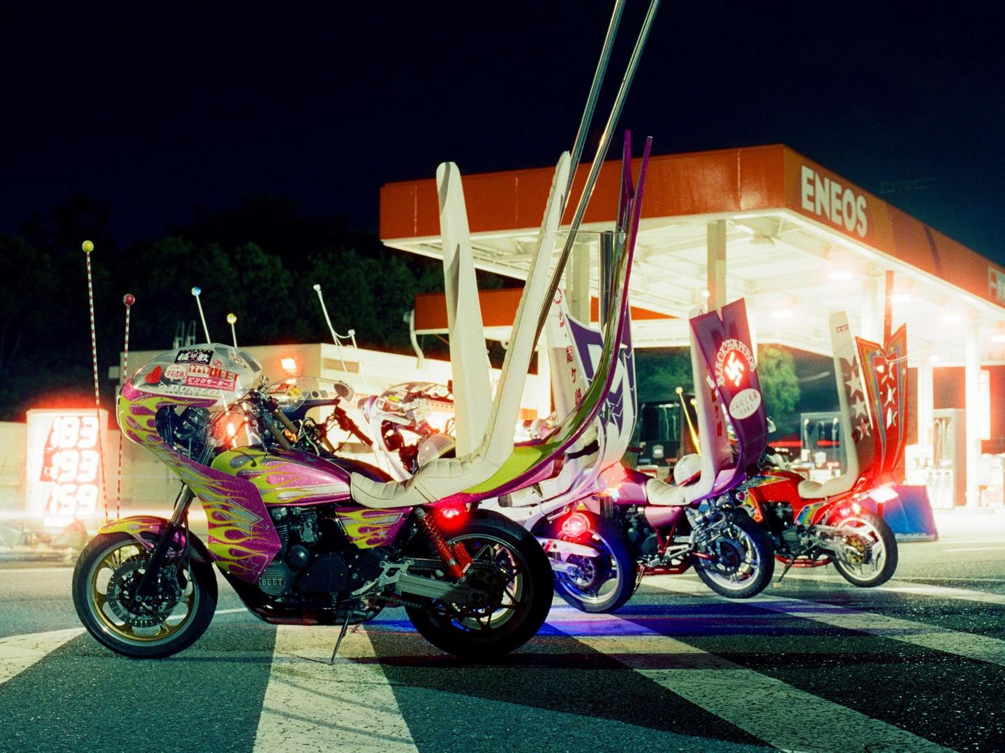 Four highly customized motorcycles with tall backrests are parked at night in front of an Eneos gas station.