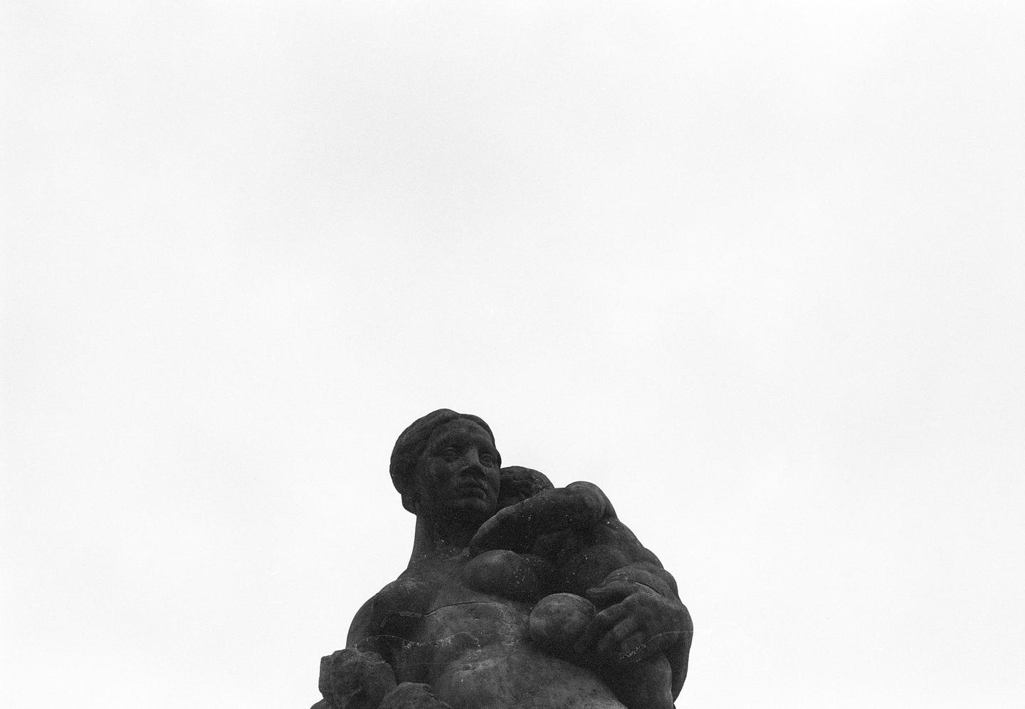 A dark statue of a woman holding a child is visible against a bright white sky.