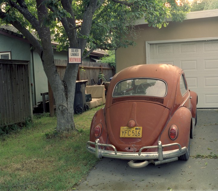 A red vintage car is parked in a driveway next to a tree with a sign that reads "Silver Linings Everywhere."
