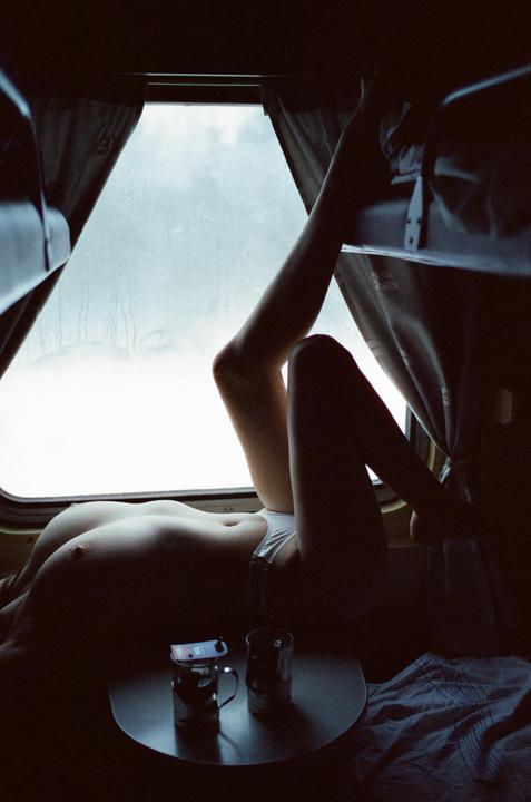 A woman lies topless on a train bunk next to a window with two cups on a table.