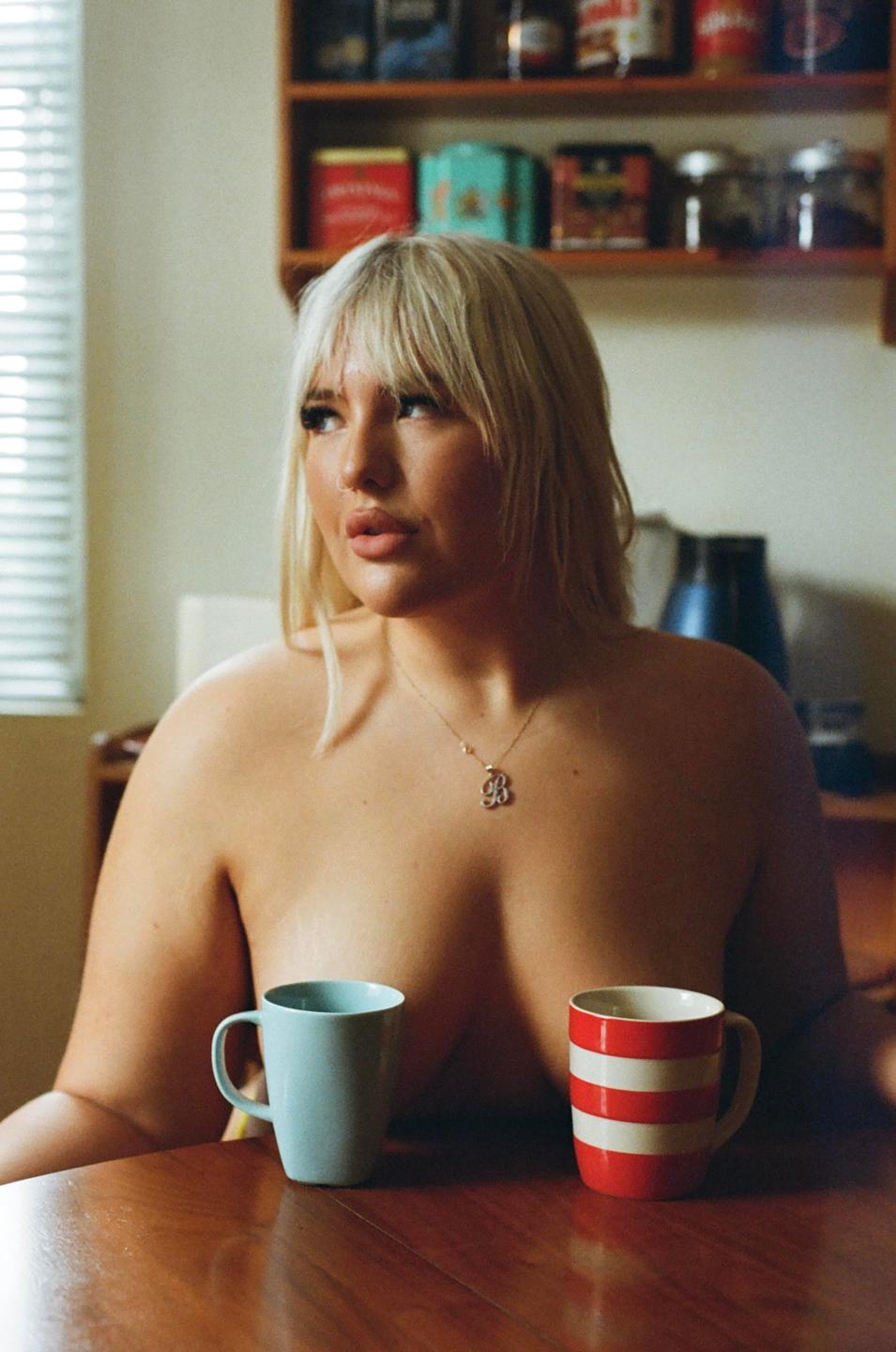 A woman with blonde hair sits at a wooden table with two mugs, looking away from the camera.