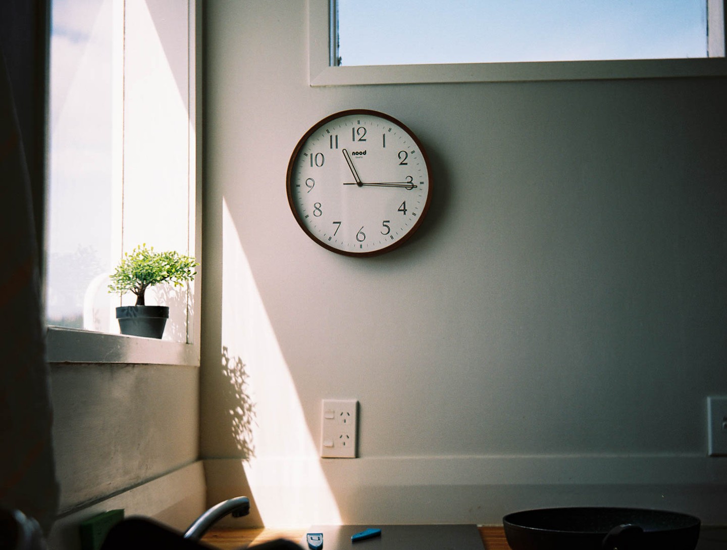Sunlight streams through a window onto a wall with a clock and a small potted plant on the windowsill.