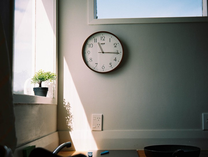 Sunlight streams through a window onto a wall with a clock and a small potted plant on the windowsill.