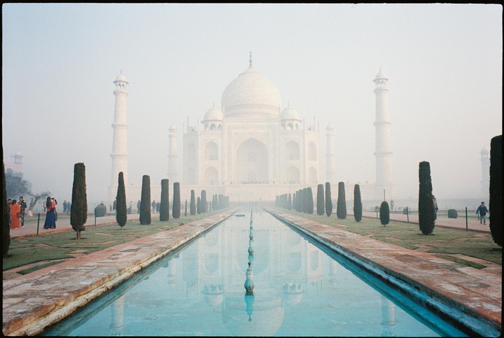 The Taj Mahal is seen through a hazy atmosphere, reflected in a long pool of water with cypress trees lining its sides.