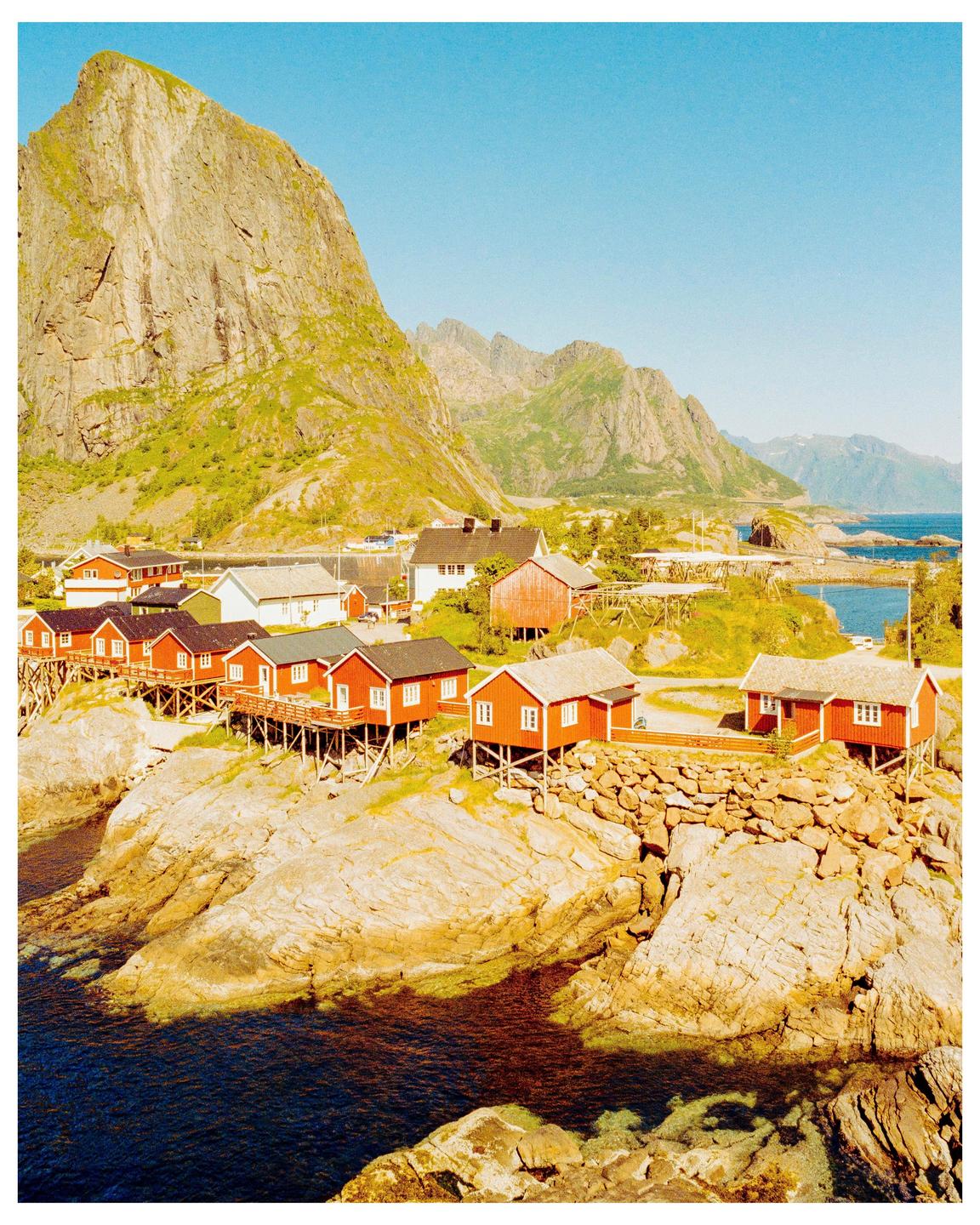 Red houses on stilts are built on a rocky coastline with a large mountain in the background.