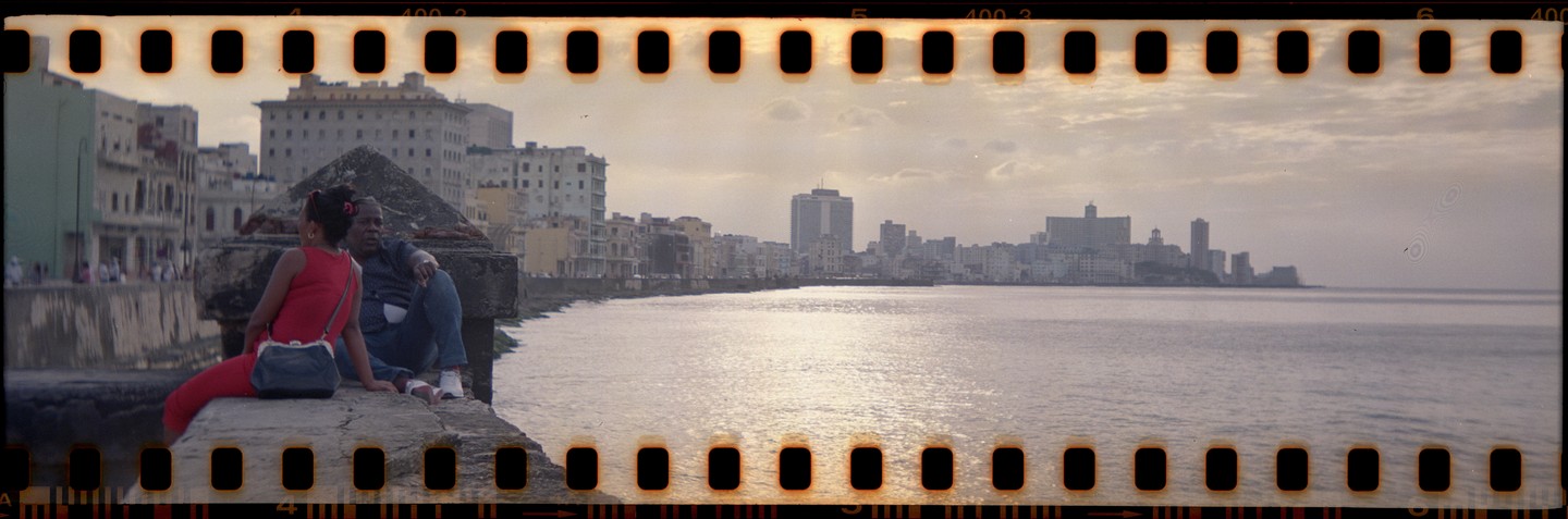 A couple sits on a stone wall overlooking a city skyline and water.