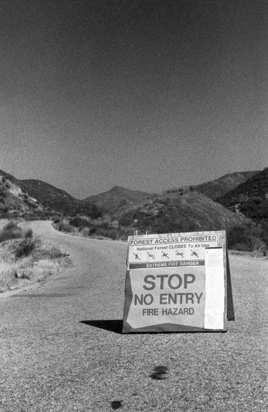 A "Stop No Entry Fire Hazard" sign stands on an asphalt road leading into a hilly, arid landscape.