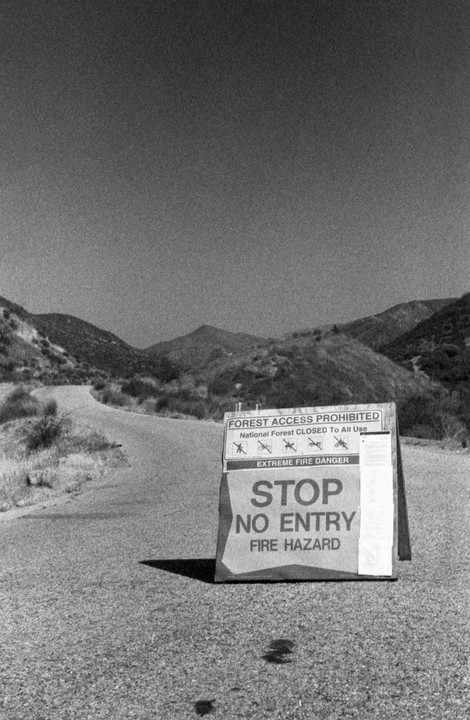 A "Stop No Entry Fire Hazard" sign stands on an asphalt road leading into a hilly, arid landscape.