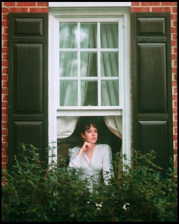 A woman looks out of a window with dark shutters and a brick wall.
