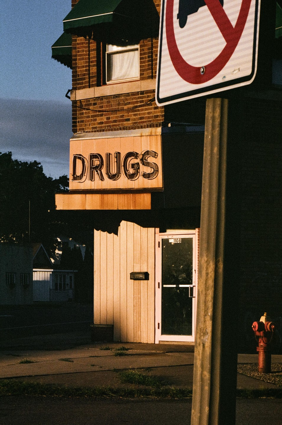 A "DRUGS" sign is visible on a brick building with a green awning, next to a street sign and a fire hydrant.