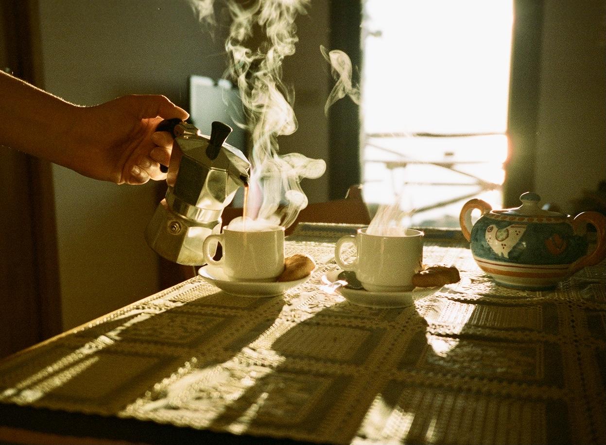 A hand pours coffee from a moka pot into a steaming cup on a table with another cup and a teapot.