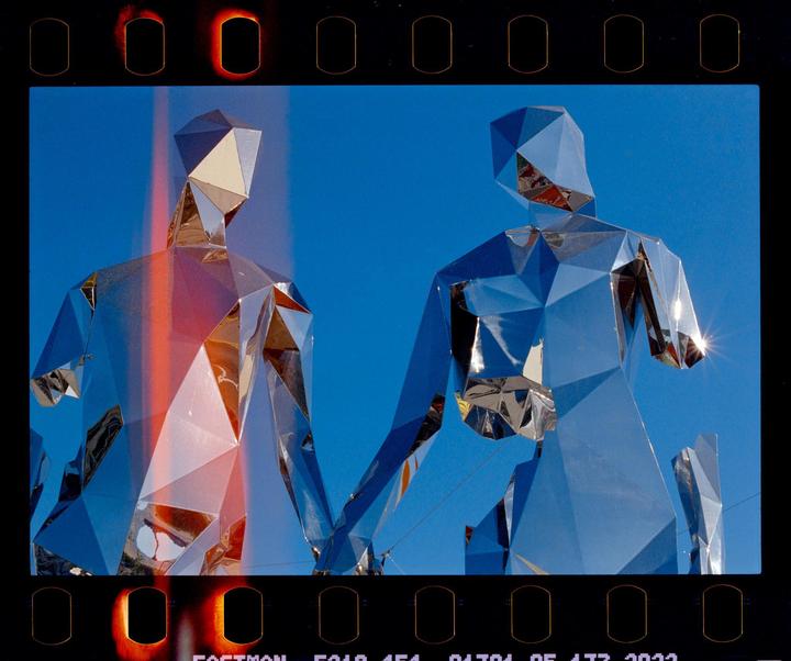 Two faceted, mirrored figures stand against a clear blue sky, with one figure showing a red light streak.