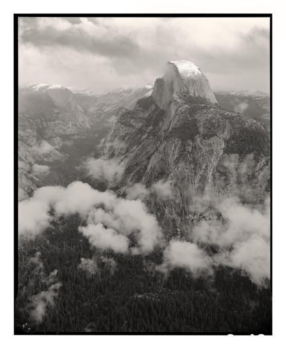 Clouds hang over a valley with a large mountain in the background and a forest in the foreground.