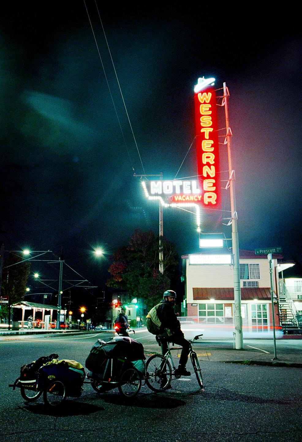 A man on a bicycle with two trailers attached is stopped at an intersection at night, with a neon motel sign in the background.