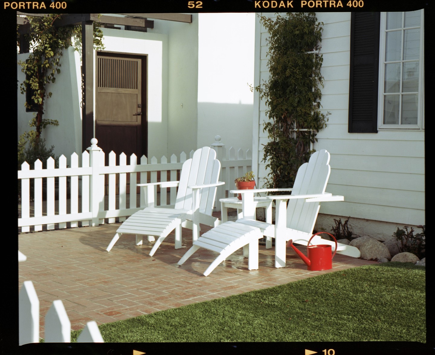 Two white Adirondack chairs and a small table sit on a brick patio next to a white picket fence and a house.