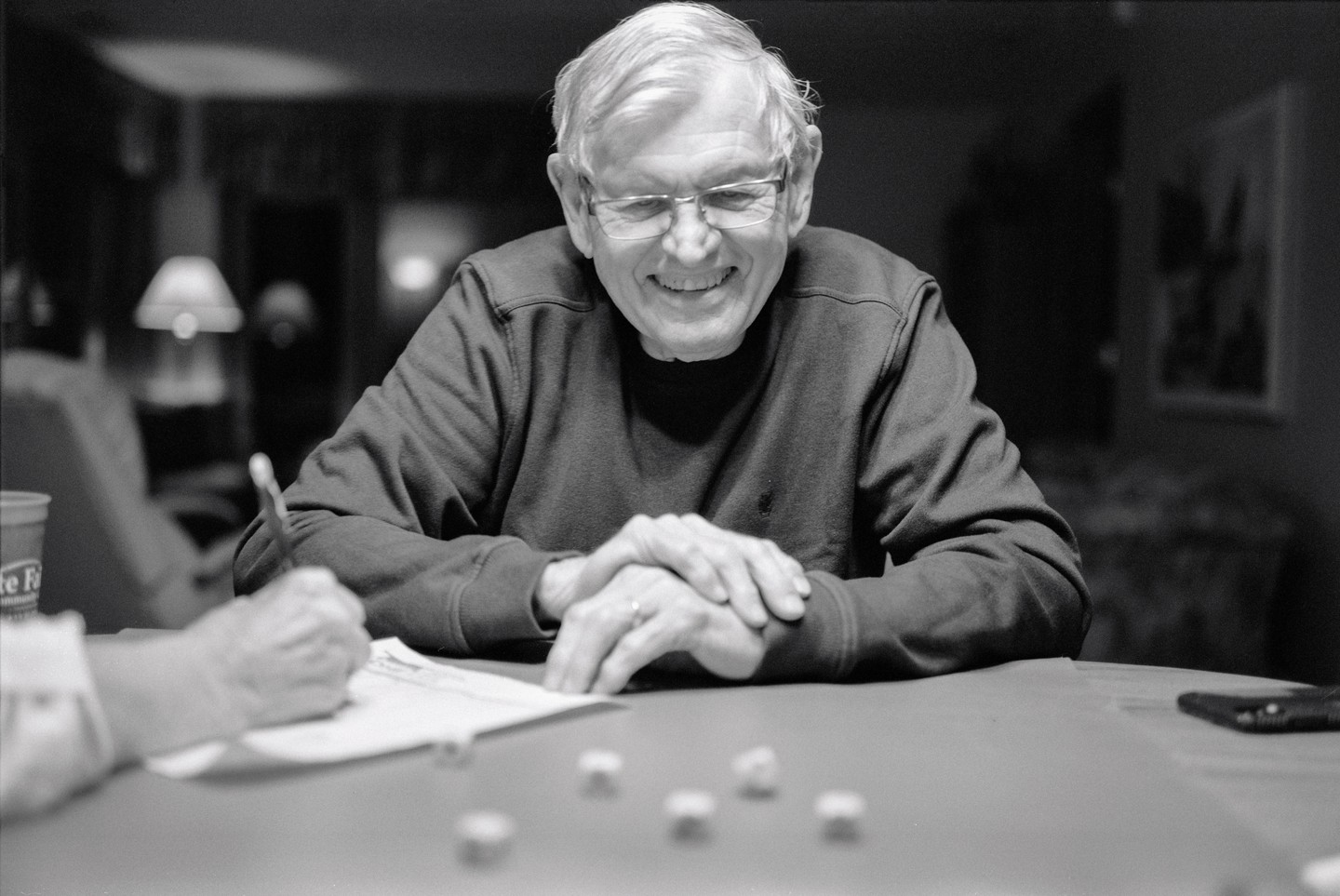 A smiling man wearing glasses sits at a table with his hands clasped, while another person's hand writes on paper next to dice.