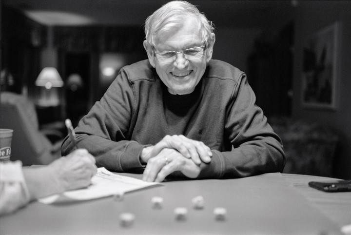A smiling man wearing glasses sits at a table with his hands clasped, while another person's hand writes on paper next to dice.