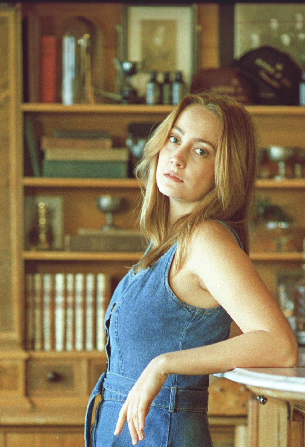 A woman in a denim dress leans on a counter with a bookshelf in the background.
