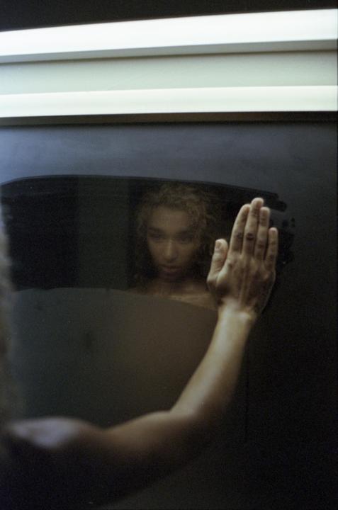 A woman with curly hair looks at her reflection in a dark surface, with her hand pressed against it.