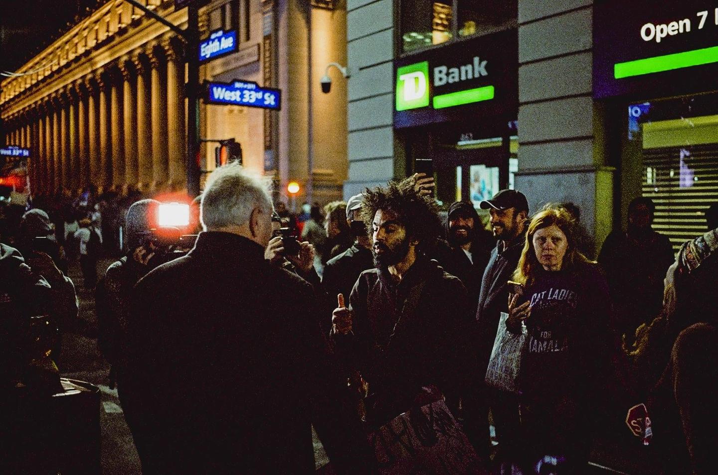 A crowd of people gathers on a city street at night, with a man in the foreground holding a camera with a bright flash.