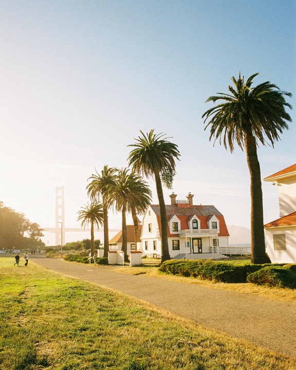 A white house with a red roof stands among palm trees, with a bridge visible in the hazy distance.