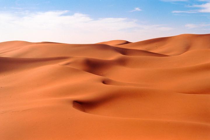 Rolling sand dunes stretch across the desert under a blue sky with scattered clouds.