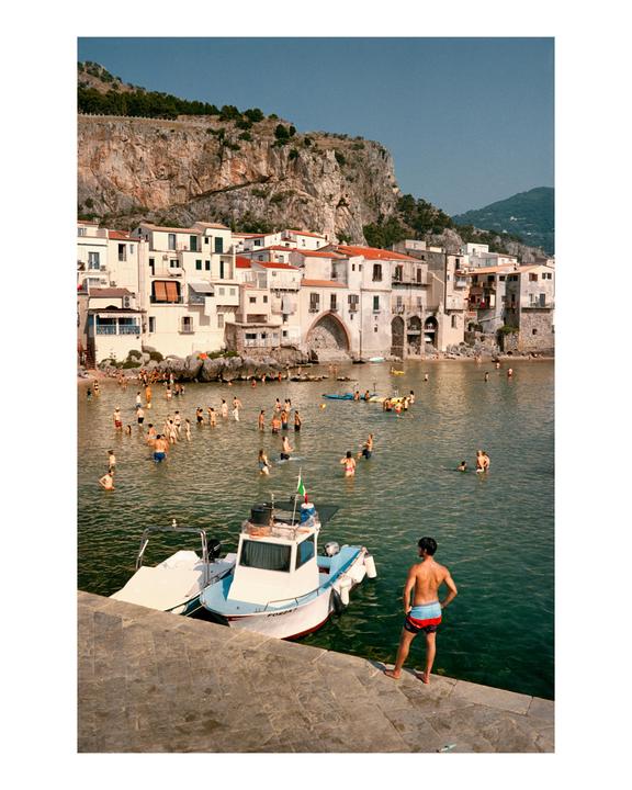 A man stands on a stone pier looking out at a bay filled with swimmers and boats, with a town built into a cliffside in the background.