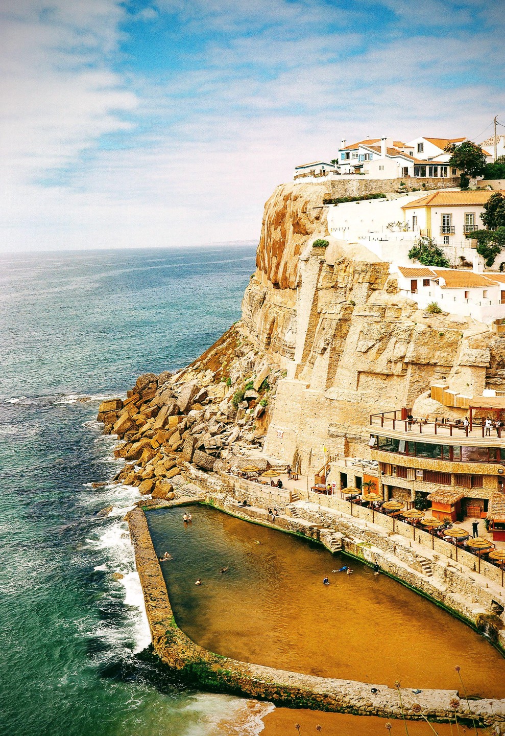 A village with white buildings and red roofs sits atop a cliff overlooking the ocean, with a natural swimming pool carved into the rocks below.