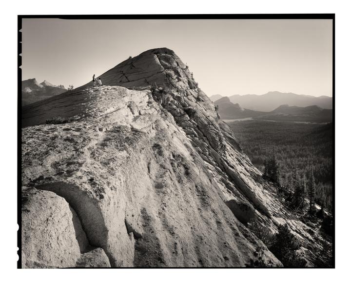 A couple stands on a rocky mountain peak overlooking a forest and distant mountains.