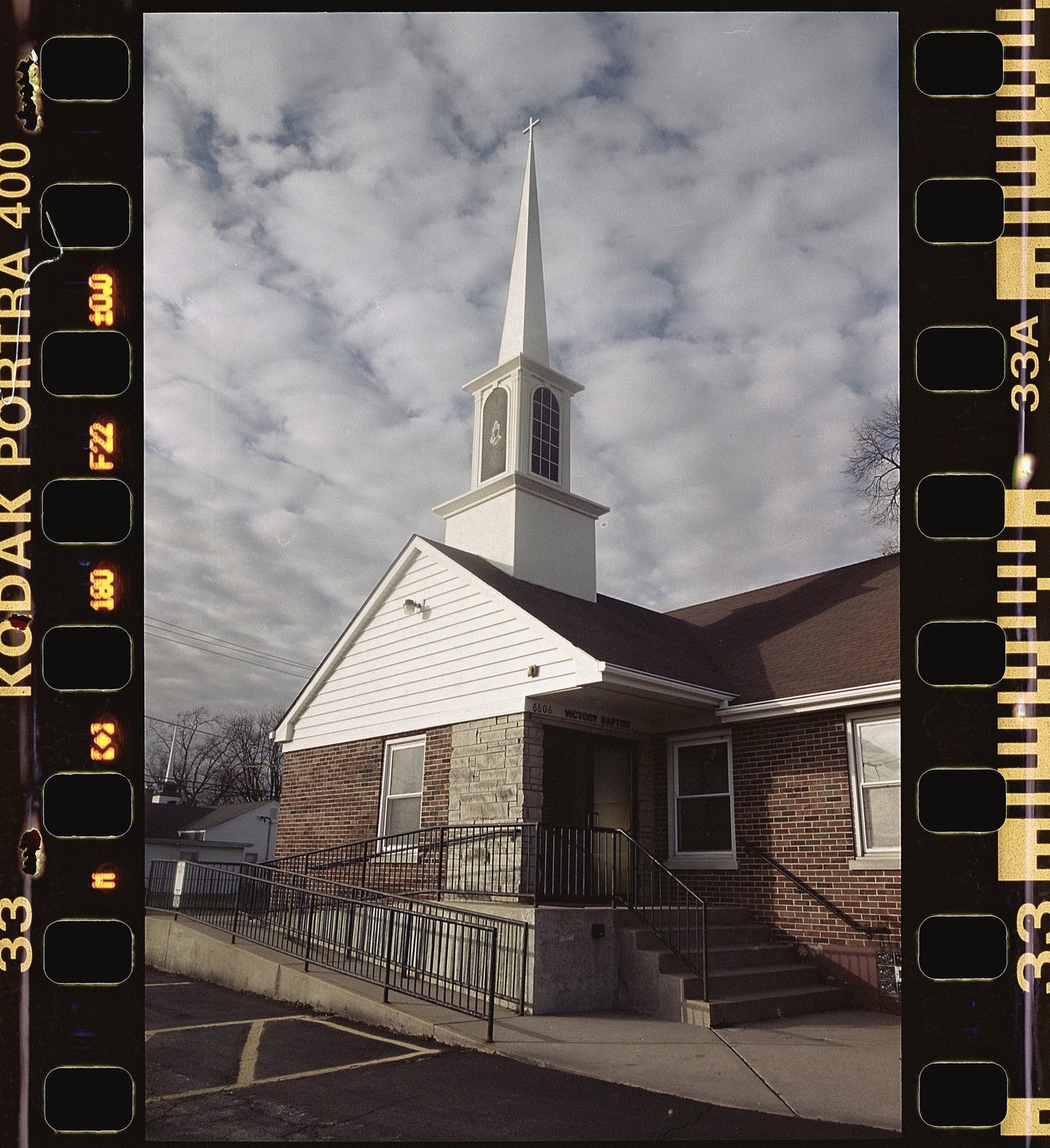 A white steeple rises above a brick and stone church with a ramp and stairs leading to its entrance under a cloudy sky.