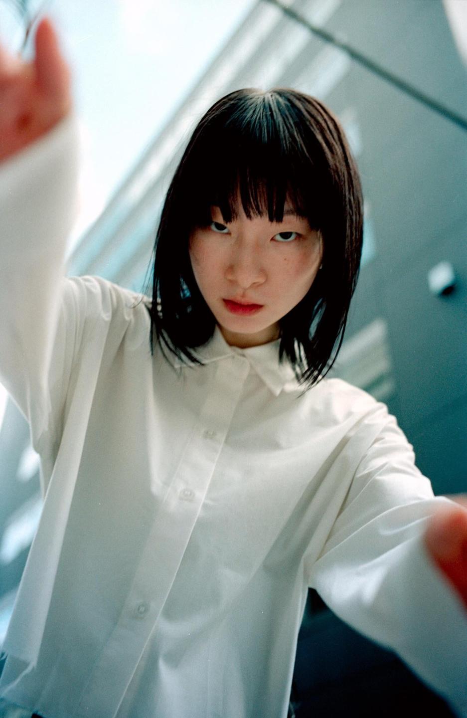 A woman with dark hair and bangs looks directly at the viewer, holding her hands up as if taking a selfie.