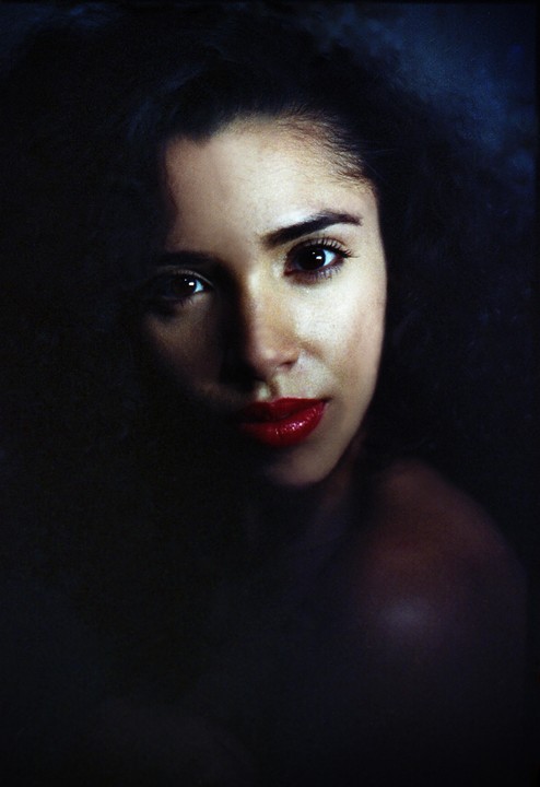 A woman with dark hair and red lipstick looks directly at the viewer, with half of her face in shadow.