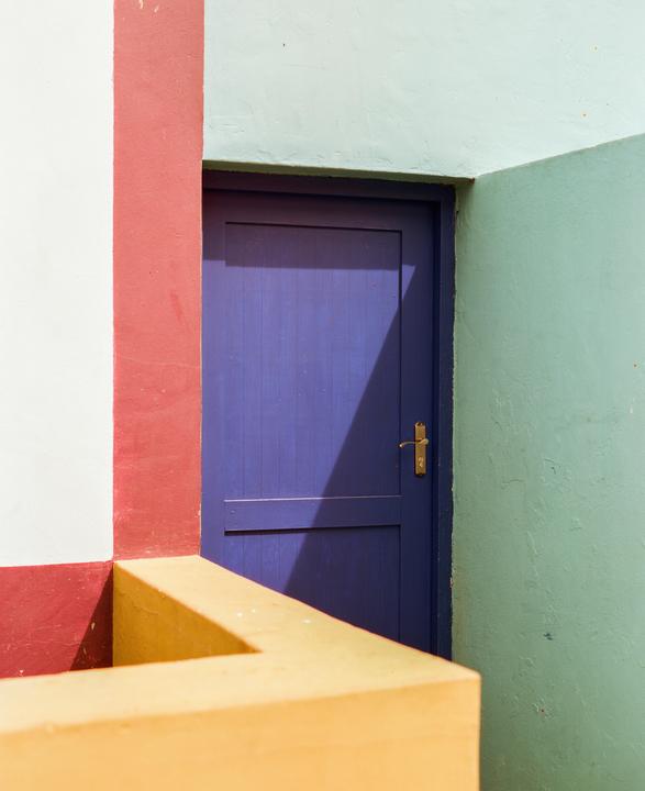 A purple door is set into a wall with red, white, and mint green sections, with a yellow railing in the foreground.