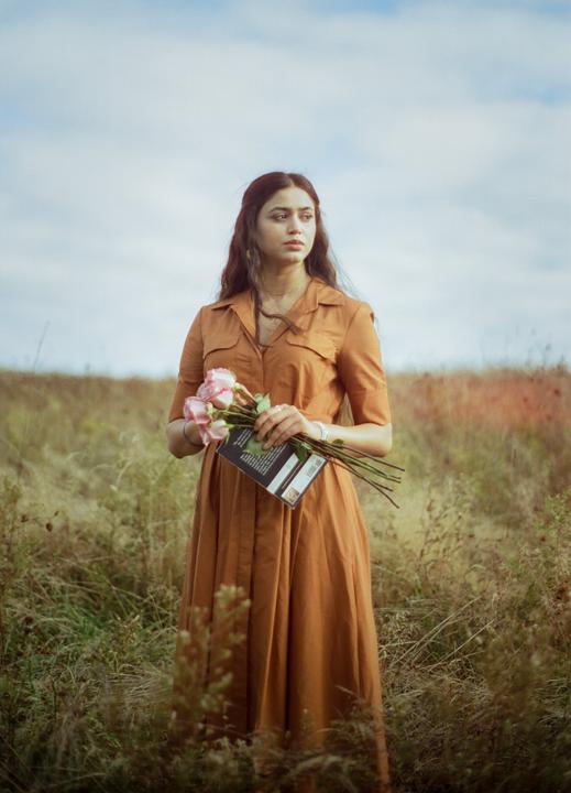A woman in a brown dress stands in a field holding a book and a bouquet of pink roses.