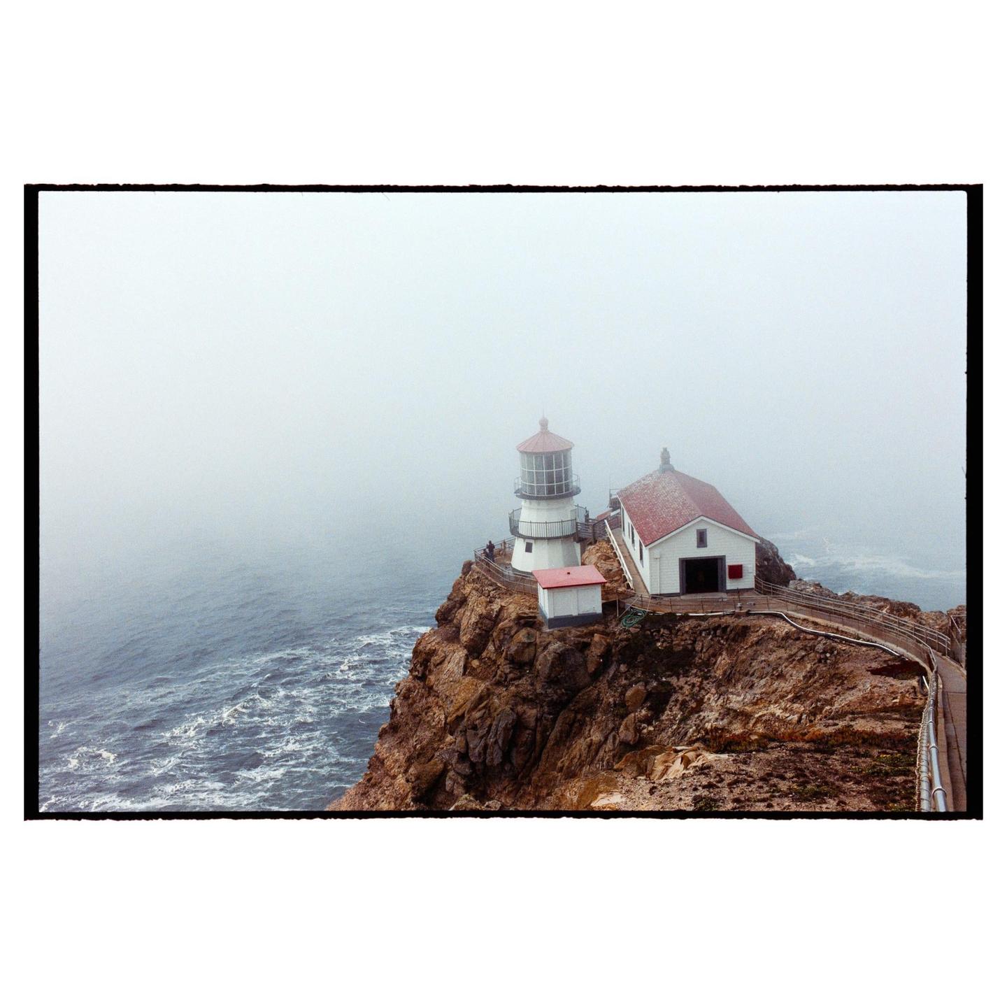 A lighthouse and building with a red roof stand on a rocky cliff overlooking the ocean on a foggy day.