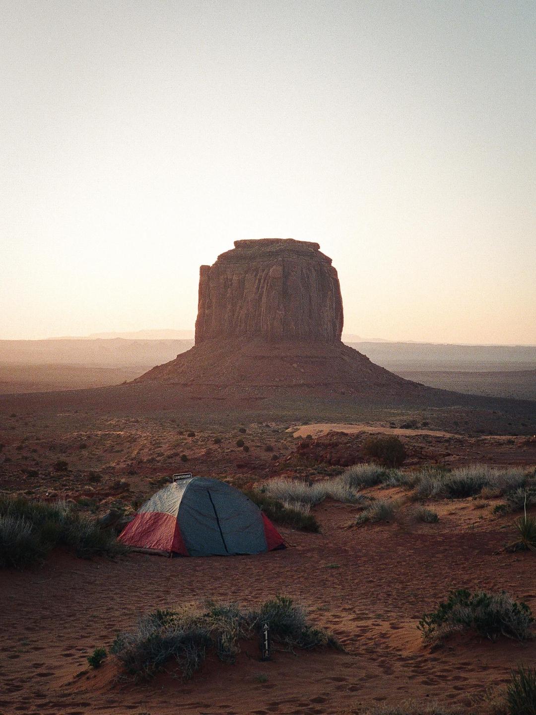 A red and gray tent is pitched in a desert landscape with a large mesa in the background.