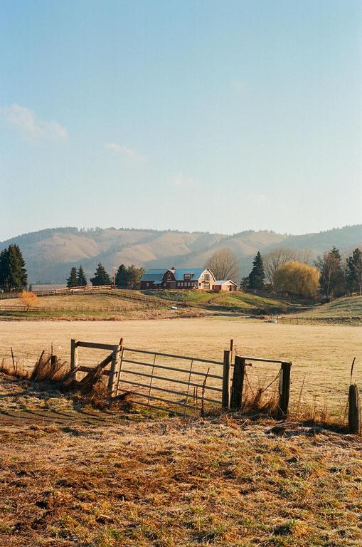 A red barn and house sit on a hill overlooking a field with a metal gate in the foreground.