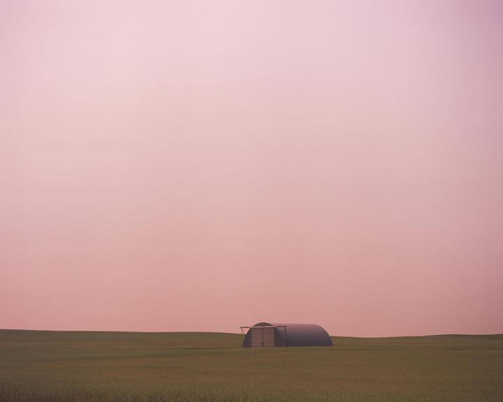 A small, dark quonset hut sits in a green field under a pink sky.