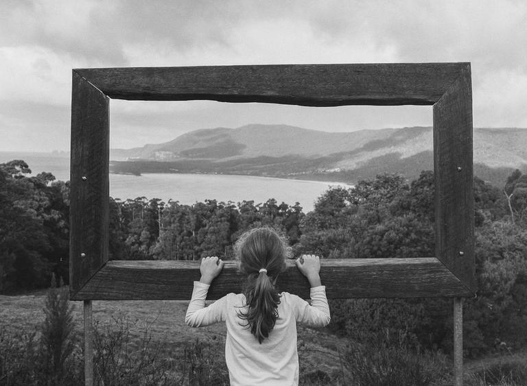 A girl looks through a large wooden frame at a bay surrounded by mountains and trees.