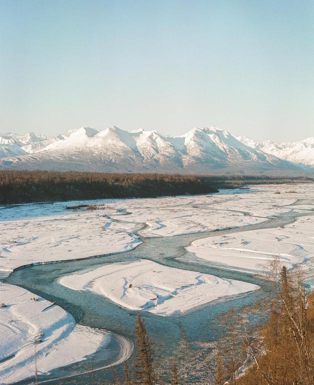 A wide river with snow-covered banks flows through a valley with a forest and snow-capped mountains in the background.