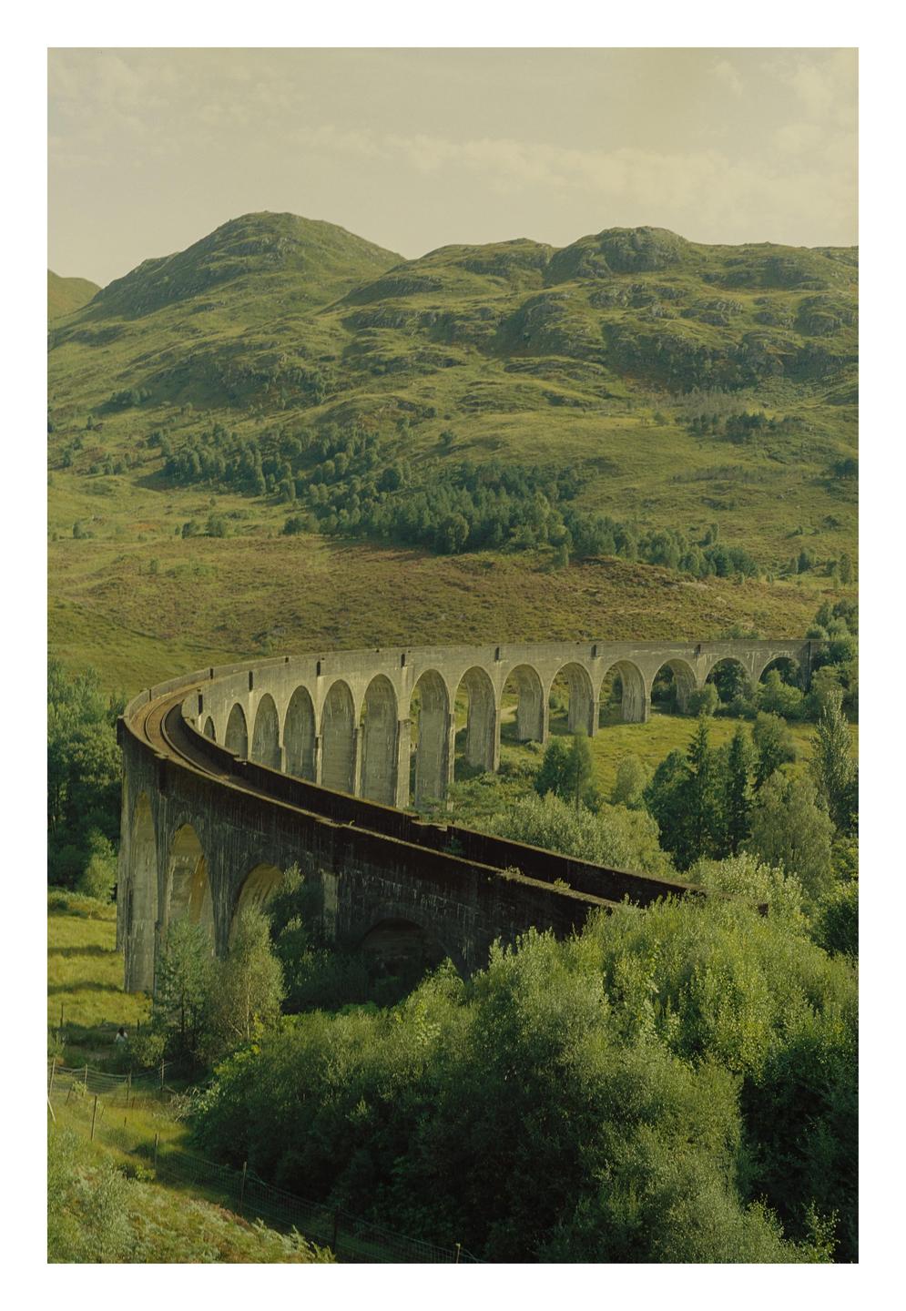 A long, curving railway viaduct with many arches spans a green valley with hills in the background.