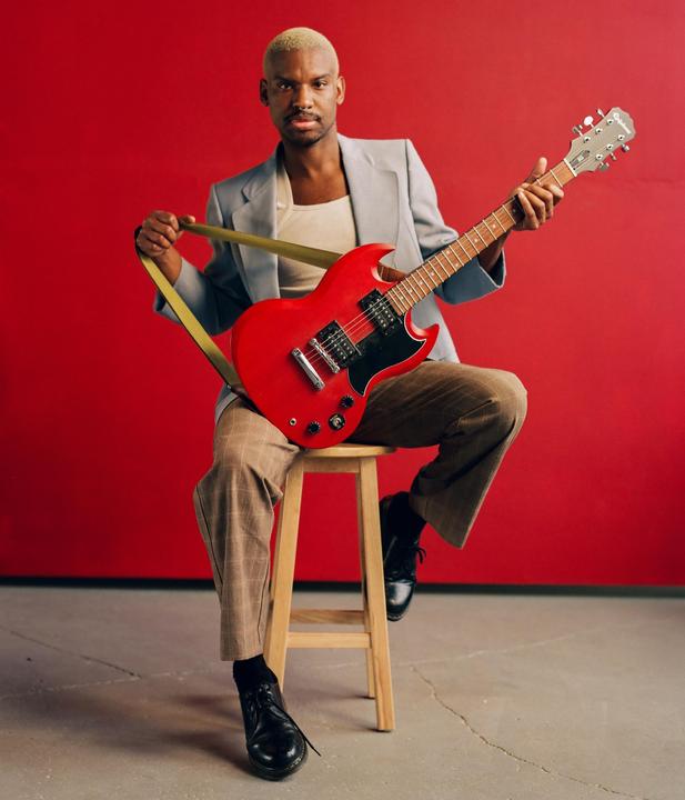 A man with bleached hair sits on a stool holding a red electric guitar against a red background.