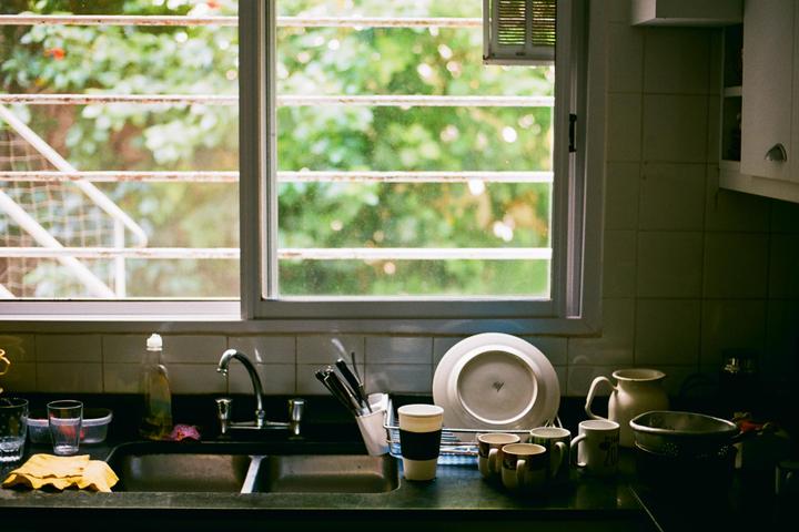 A kitchen sink and counter are filled with dishes and utensils next to a window looking out onto green foliage.