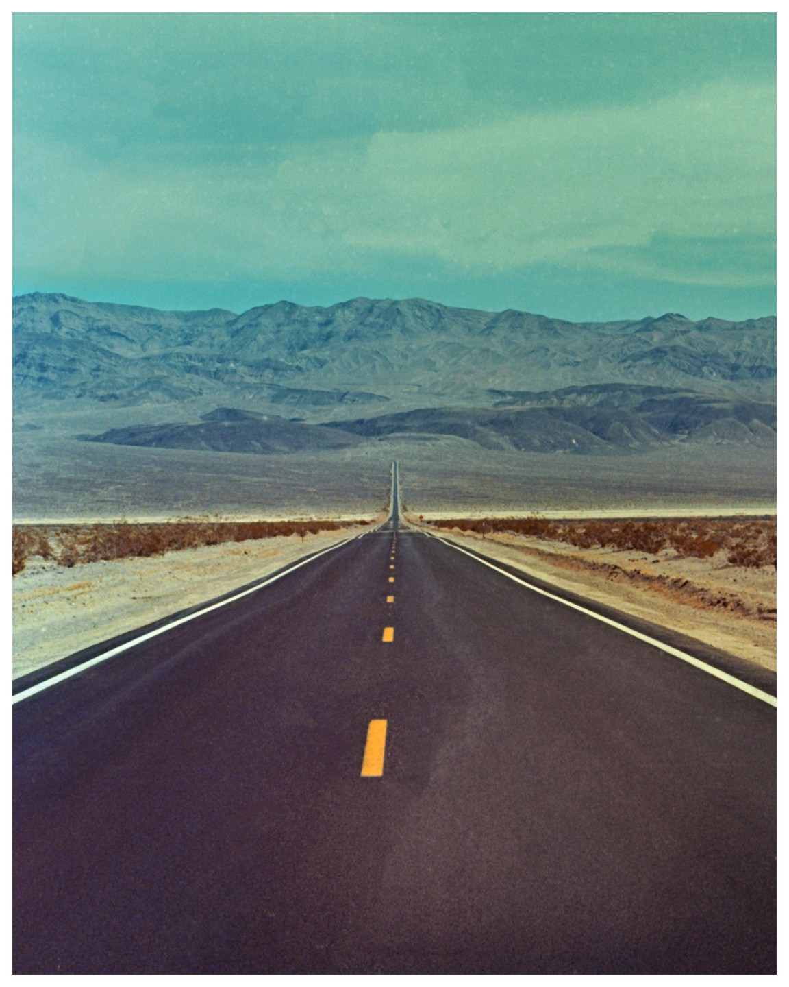 A long, straight road extends into the distance through a desert landscape with mountains under a cloudy sky.