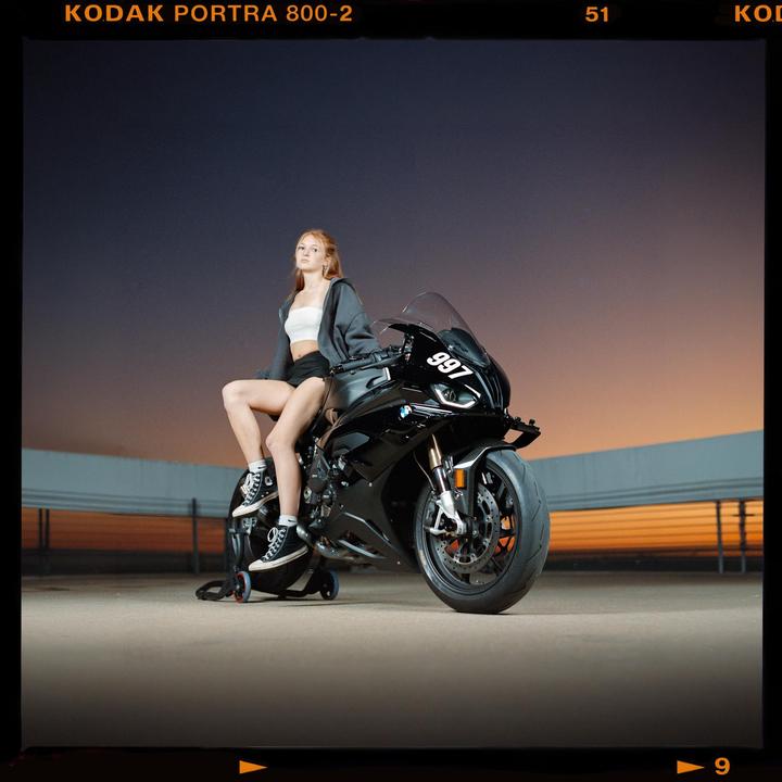 A woman sits on a black motorcycle against a sunset sky.
