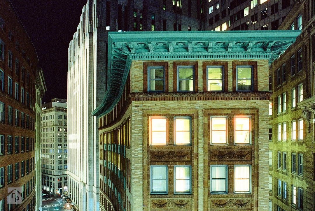 A street is visible between tall buildings with many illuminated windows at night.