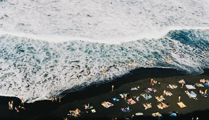 An aerial view shows people swimming in the ocean and sunbathing on a black sand beach.