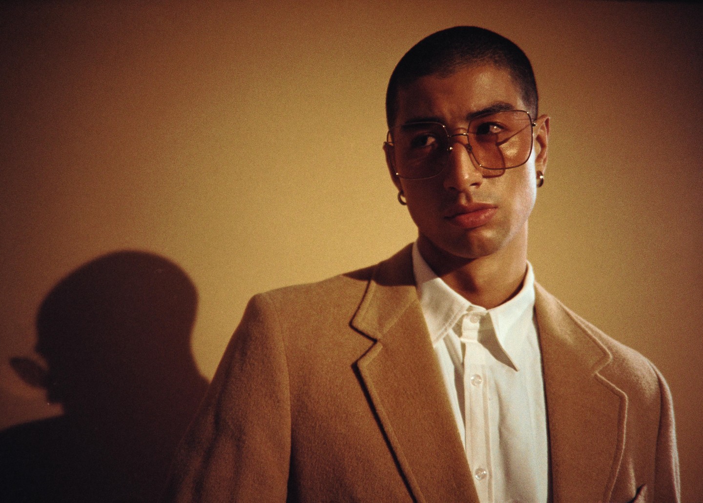 A man wearing a tan jacket and glasses looks to the side, with a shadow of his head visible on the wall behind him.